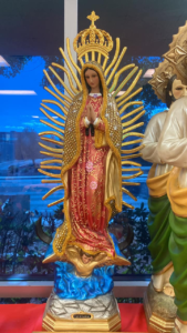 A colorful statue of the Virgin of Guadalupe with a gold crown and rays, standing on a blue cloud base